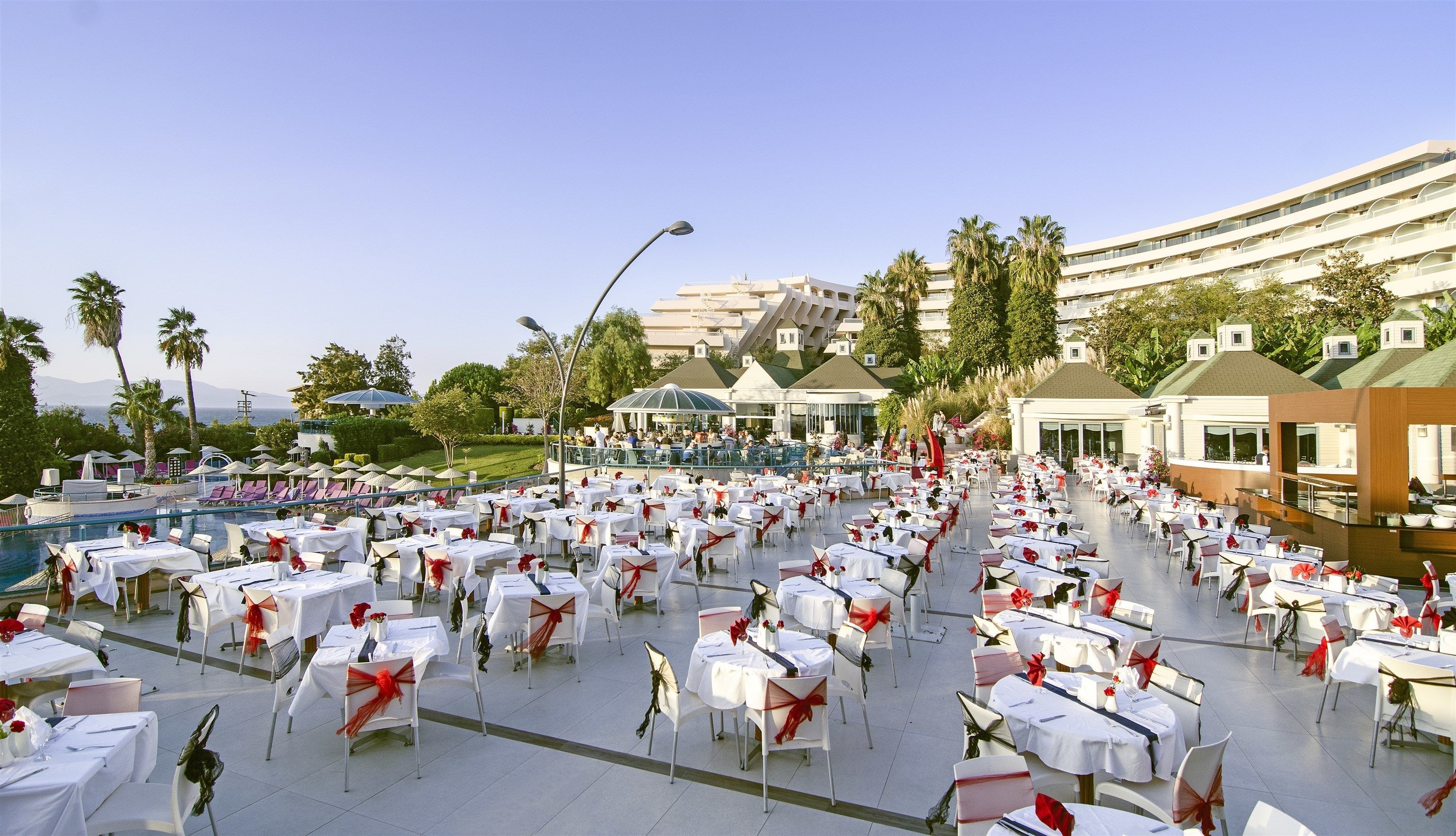 Un espacio de comedor al aire libre con numerosas mesas redondas, cada una con mantel blanco y fajín rojo.