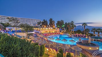 Un área de piscina en un resort, con múltiples sillas de descanso y sombrillas, rodeada de palmeras y un edificio grande al fondo.