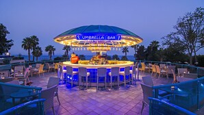 An outdoor bar with a neon sign, bar stools, and a view of the ocean. - The Grand Blue Sky International (Kusadasi)