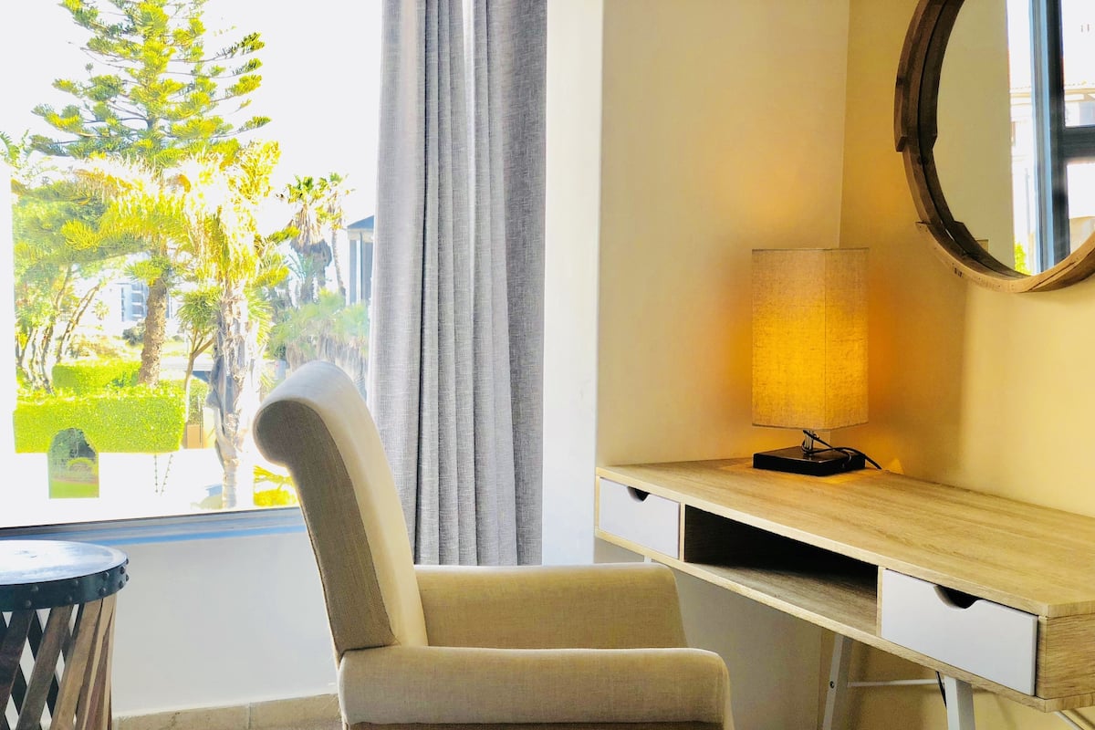 A room with a chair, a desk, a lamp, and a window with a view of greenery and buildings.