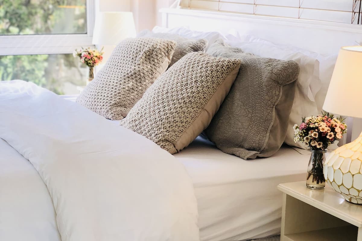 A neatly made bed with white and beige bedding, a small bedside table with a vase of flowers, and a window with blinds.