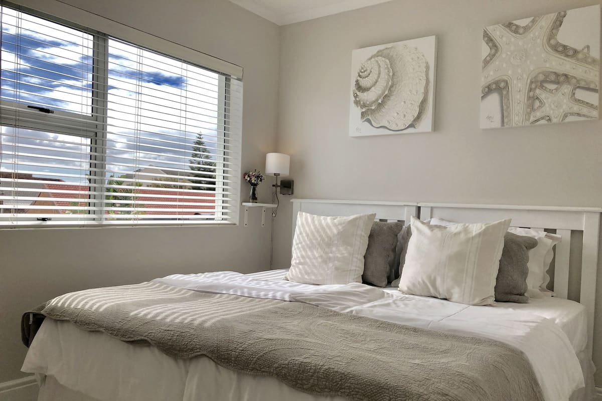 A bedroom with a bed, white bedding, a grey blanket, and a window with blinds.
