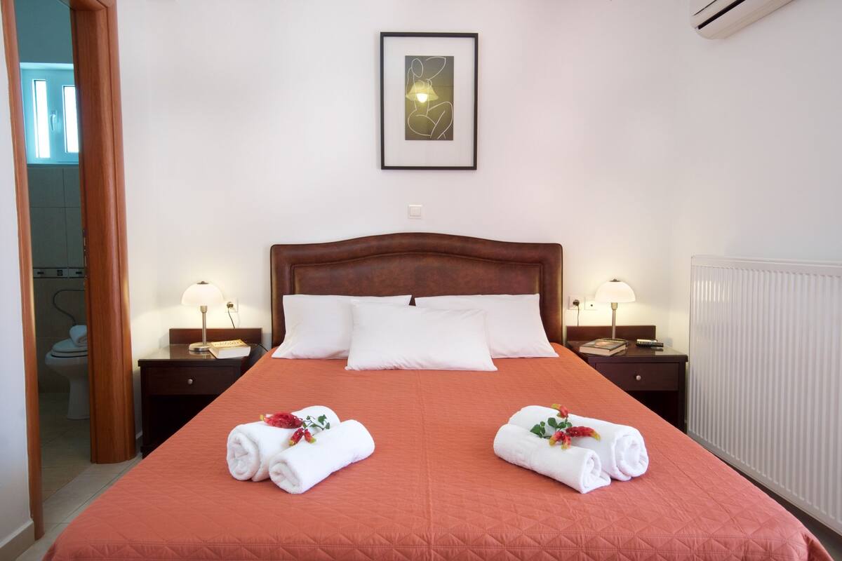 A neatly made bed with a red quilt, two bedside tables with lamps, a framed picture on the wall, and a radiator.