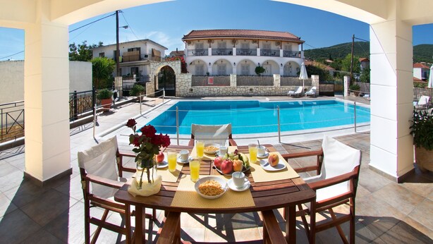 A dining area with a pool view, featuring a table set with breakfast, a stone wall, and a building in the background. - Studios Diamanto (Kefalonia)