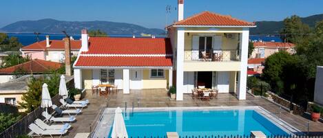 A two-story building with a swimming pool, lounge chairs, and a view of the sea.