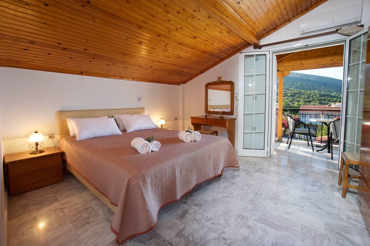 A bedroom with a large bed, wooden ceiling, a nightstand, a dresser, a mirror, and a balcony with a view of the mountains.