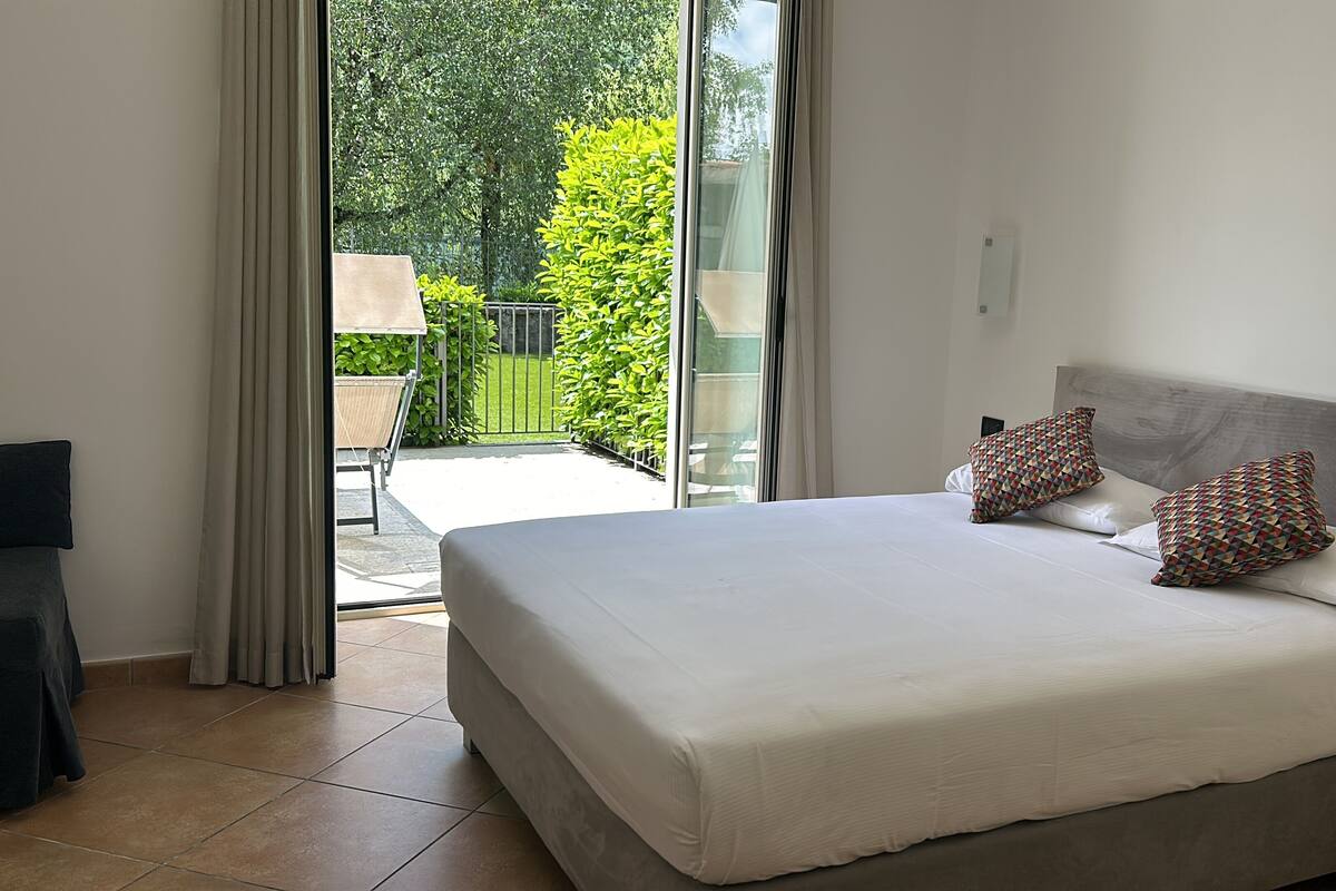 A bedroom with a large bed, two patterned pillows, a nightstand, and a view of a garden through a sliding door.