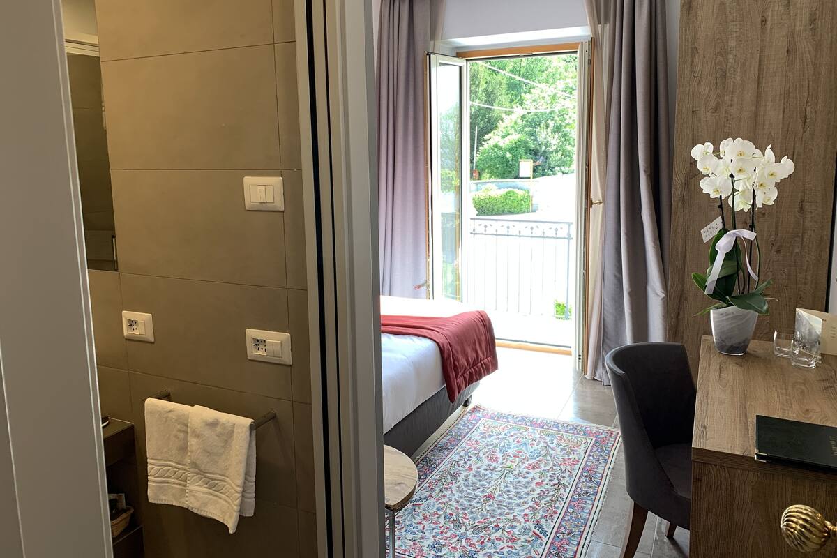 A hotel room with a bed, a bathroom with a towel rack, a desk with a vase of flowers, and a view of the outside.