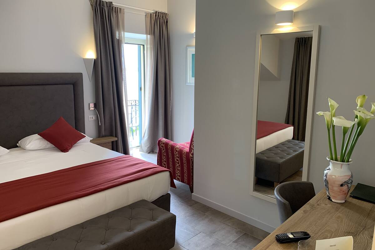 A hotel room with a large bed, a bedside table with a vase of flowers, a chair, and a desk with a mirror.
