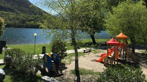 Un parque infantil con toboganes y estructuras de escalada cerca de un lago, rodeado de árboles y un banco.