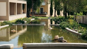 A tranquil courtyard with a small pond, surrounded by greenery and a building with large windows. - Folies Corfu Hotel Apartments (Corfu)