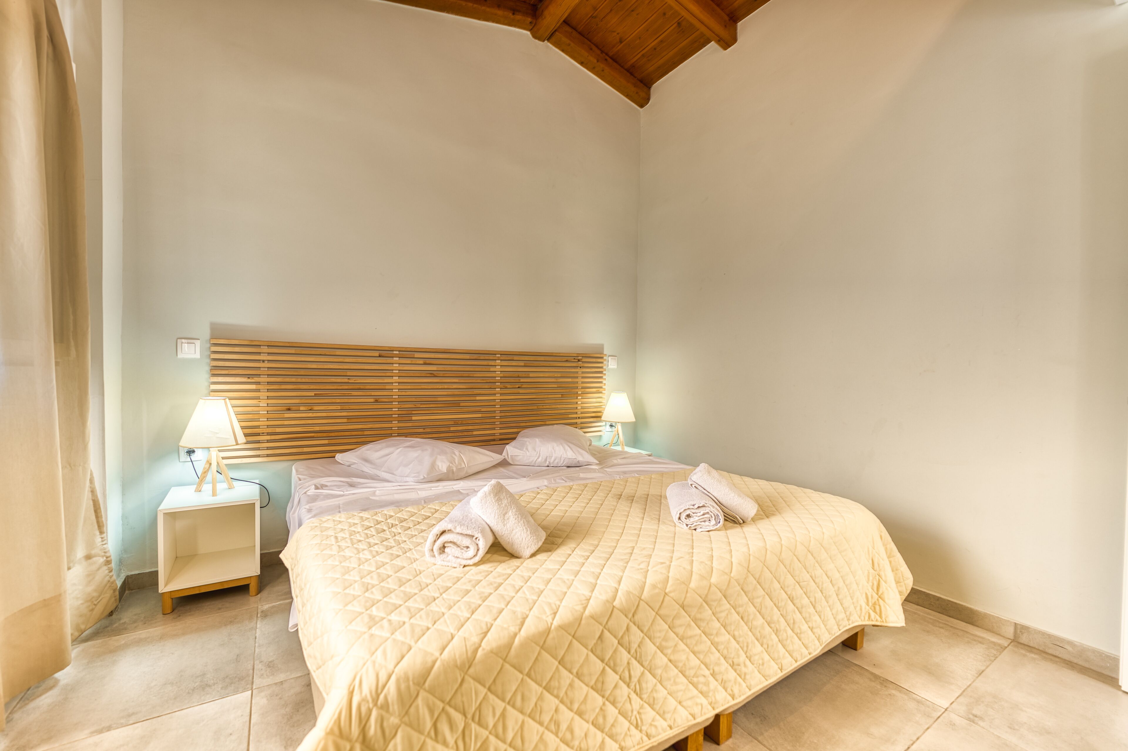Een net opgemaakt bed met een gewatteerde sprei en twee opgerolde handdoeken, een nachtkastje met een lamp, en een houten hoofdbord.