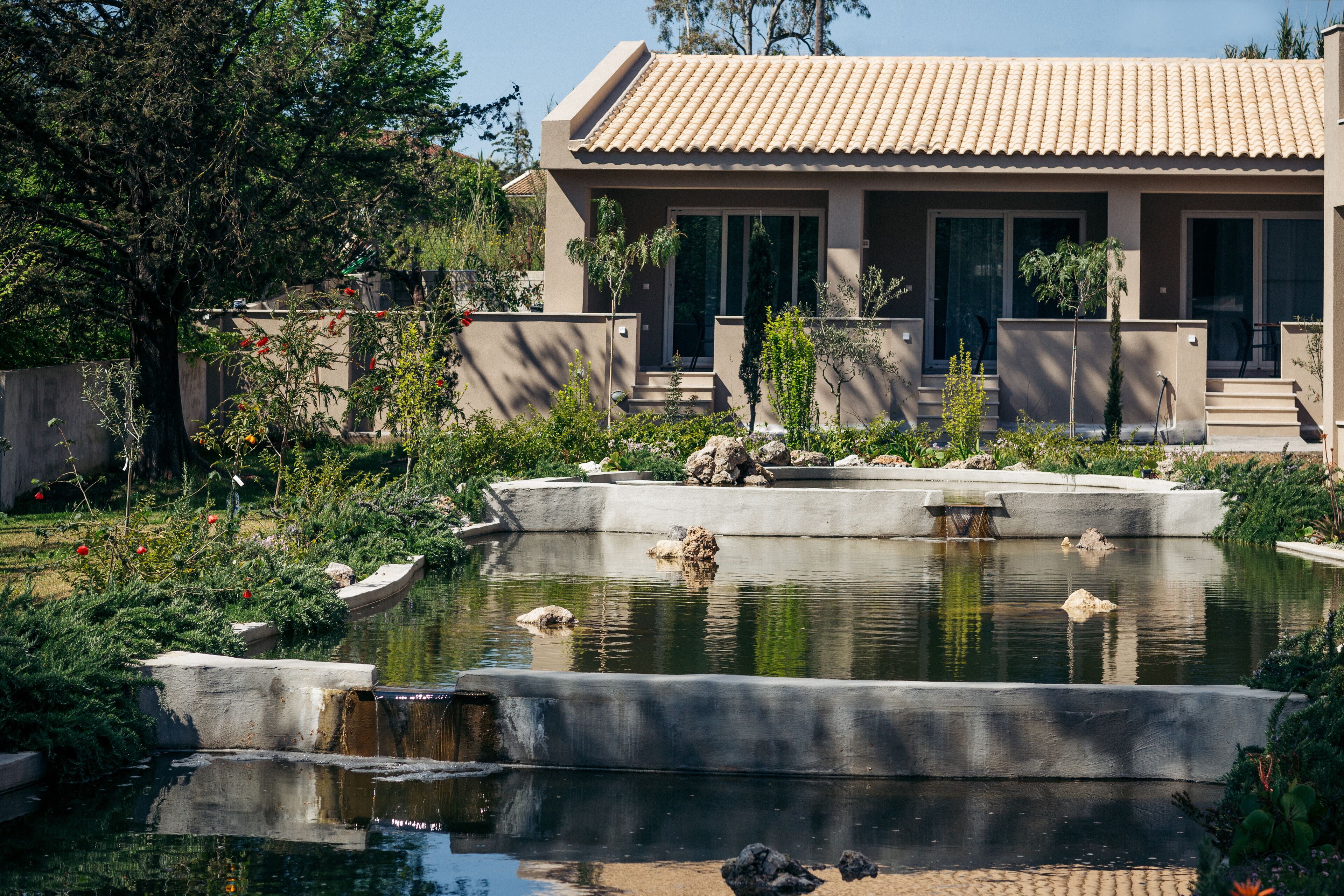 Une maison moderne avec un toit en tuiles, une piscine et un jardin agrémenté de diverses plantes et d’un petit bateau.