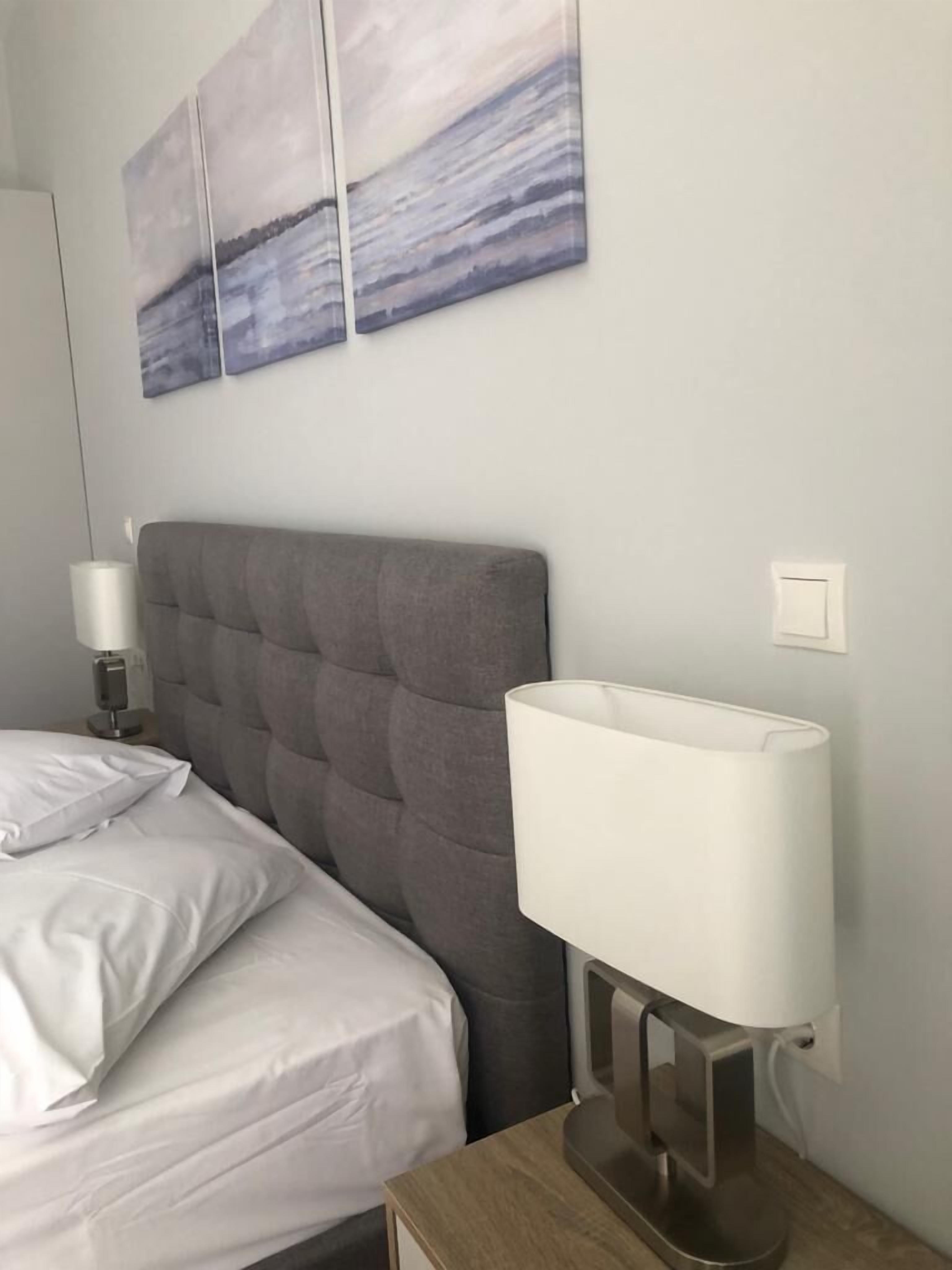 Un lit bien fait, avec du linge de lit blanc, une tête de lit rembourrée de couleur grise, une lampe de chevet et un tableau accroché au mur.