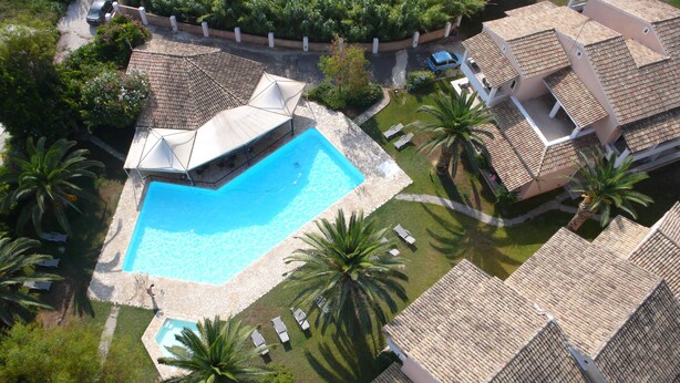 An aerial view of a residential area with a swimming pool surrounded by palm trees and a tiled-roofed building. - Folies Corfu Hotel Apartments (Corfu)