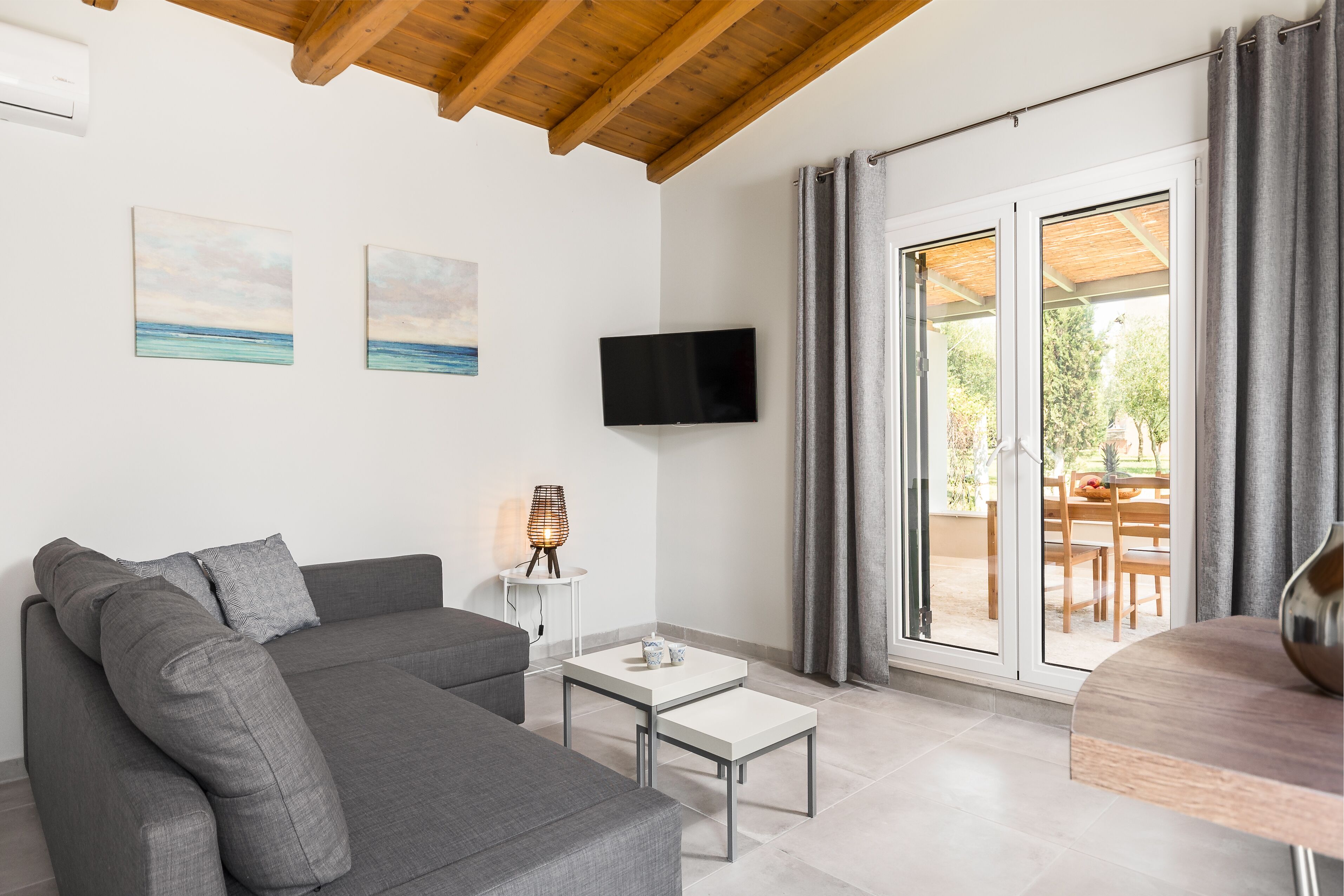 Ein modernes Wohnzimmer mit grauem Sofa, einem kleinen weißen Tisch, einem Flachbildfernseher und Blick durch Glastüren auf einen Außenbereich mit Tisch und Stühlen.