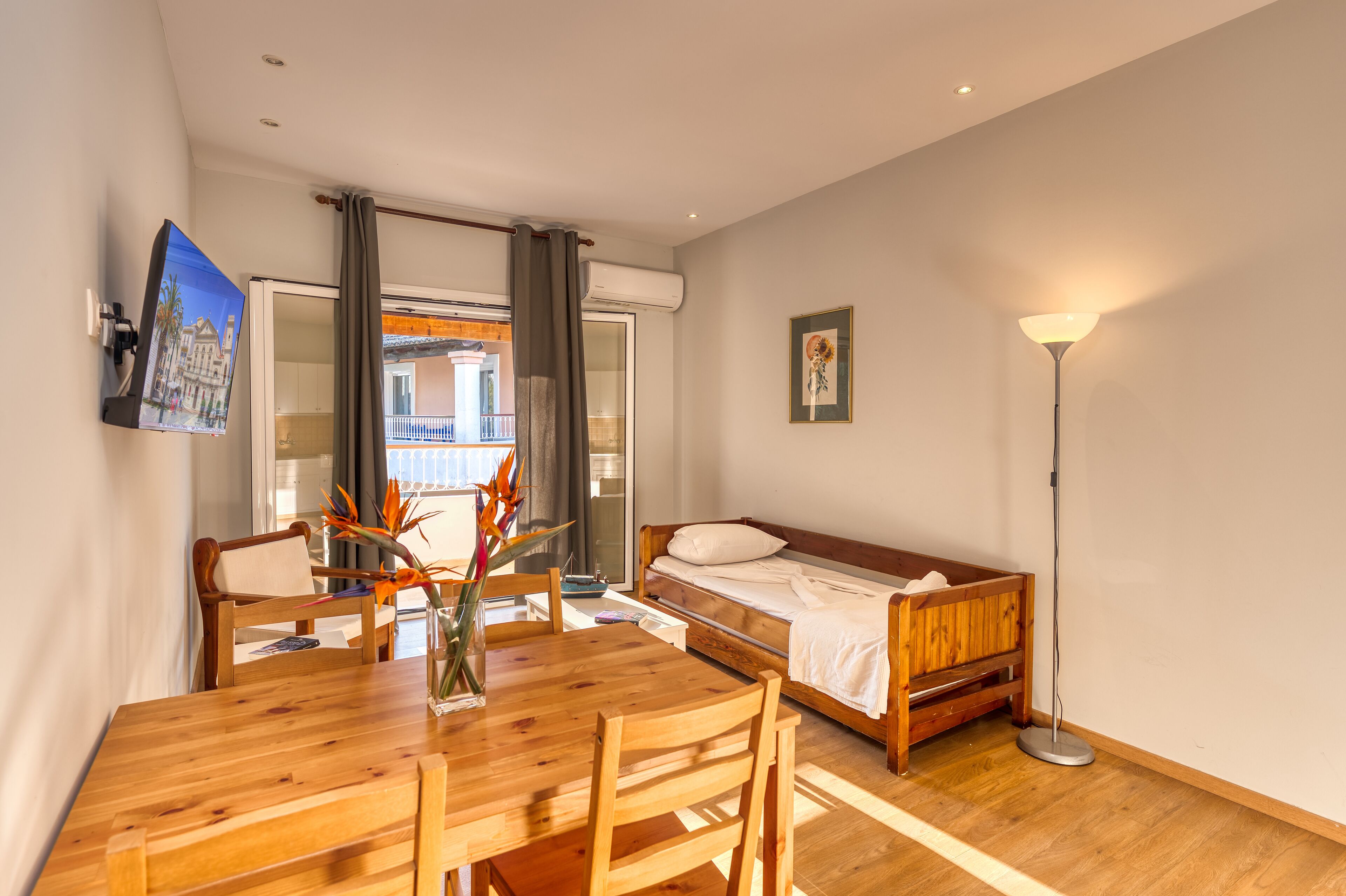 A room with a wooden dining table, a single bed, a standing lamp, and a TV mounted on the wall.