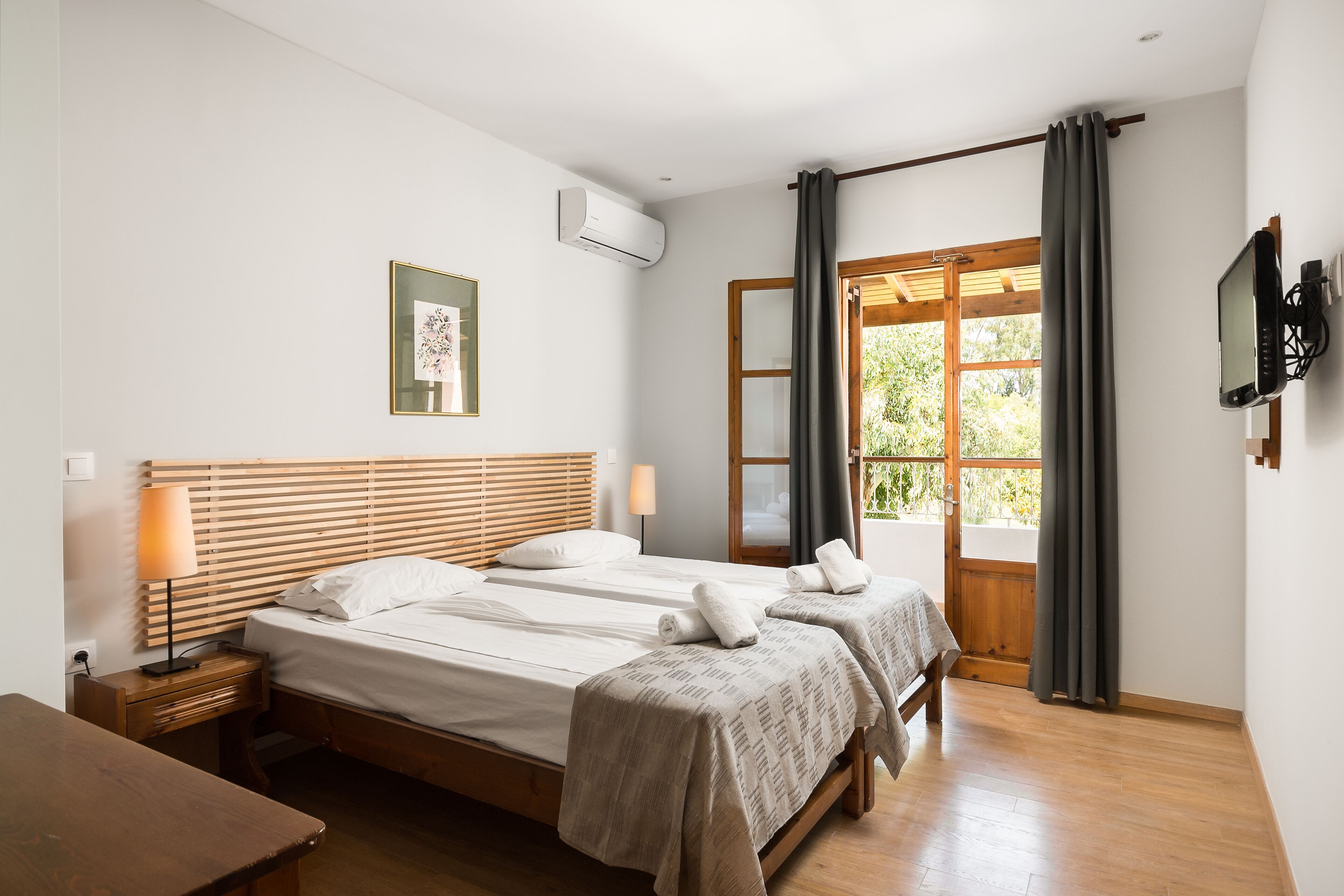 A bedroom with two beds, a wooden headboard, a bedside table with a lamp, a TV mounted on the wall, and a window with curtains.