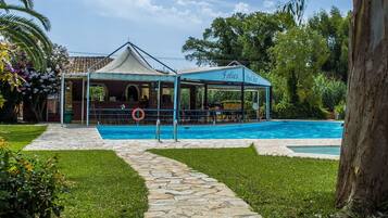Zona de piscina con bar techado y zona de descanso, sendero de piedra y exuberante vegetación.