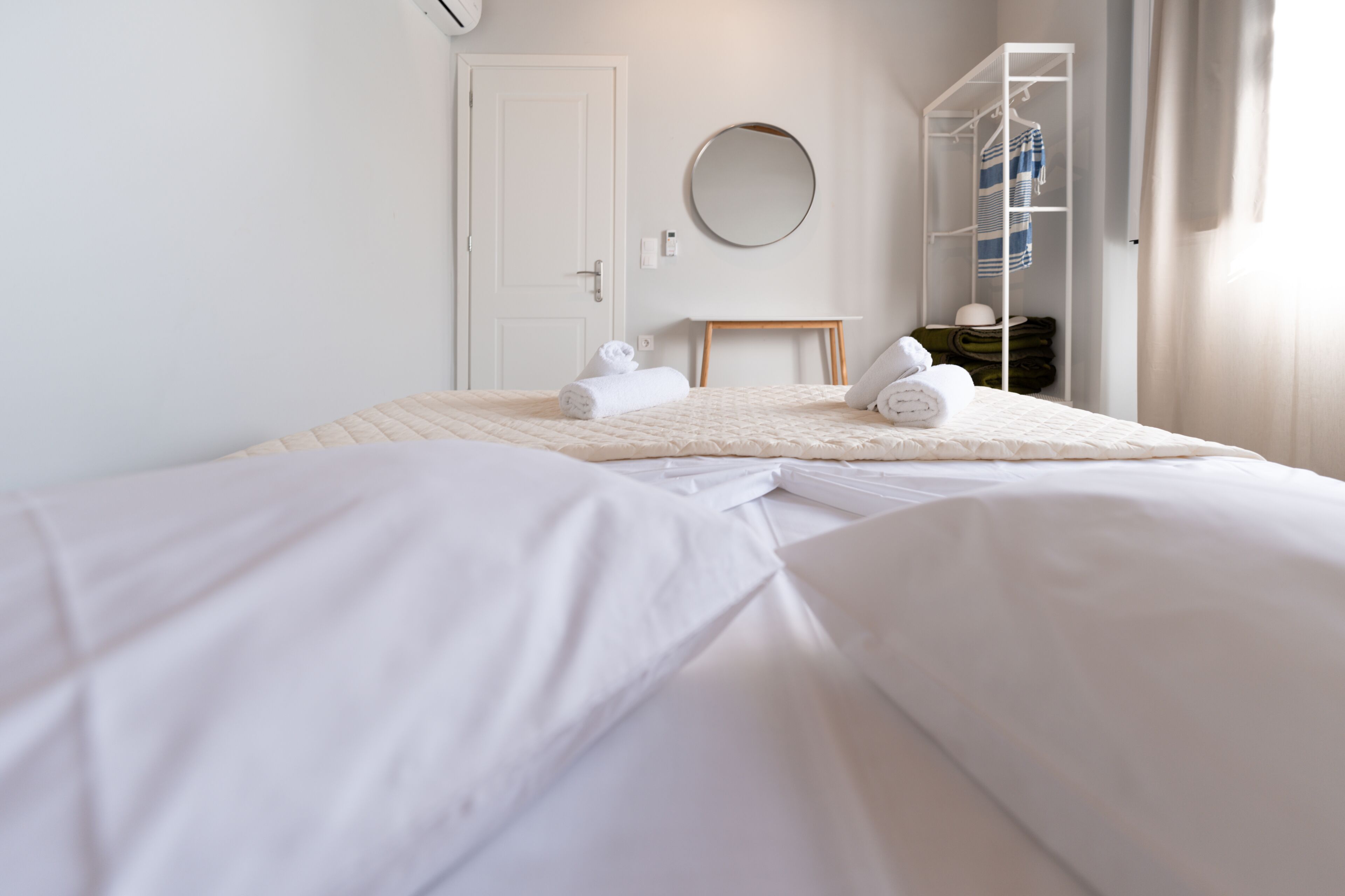 A neatly made bed with white linens, a round mirror on the wall, and a white wardrobe with a striped towel hanging.
