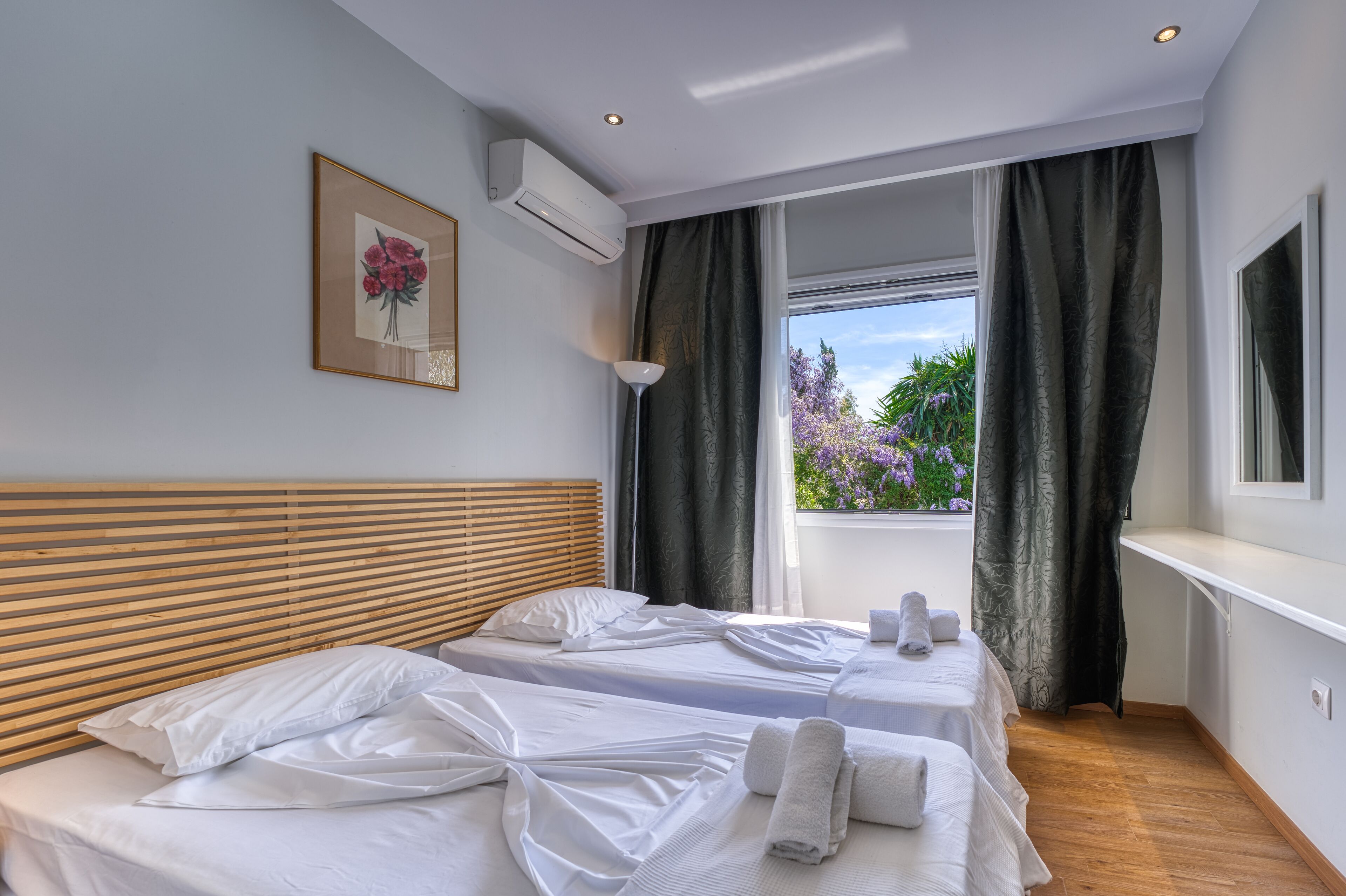 A hotel room with two beds, a wooden headboard, a window with curtains, a framed picture, and an air conditioning unit.