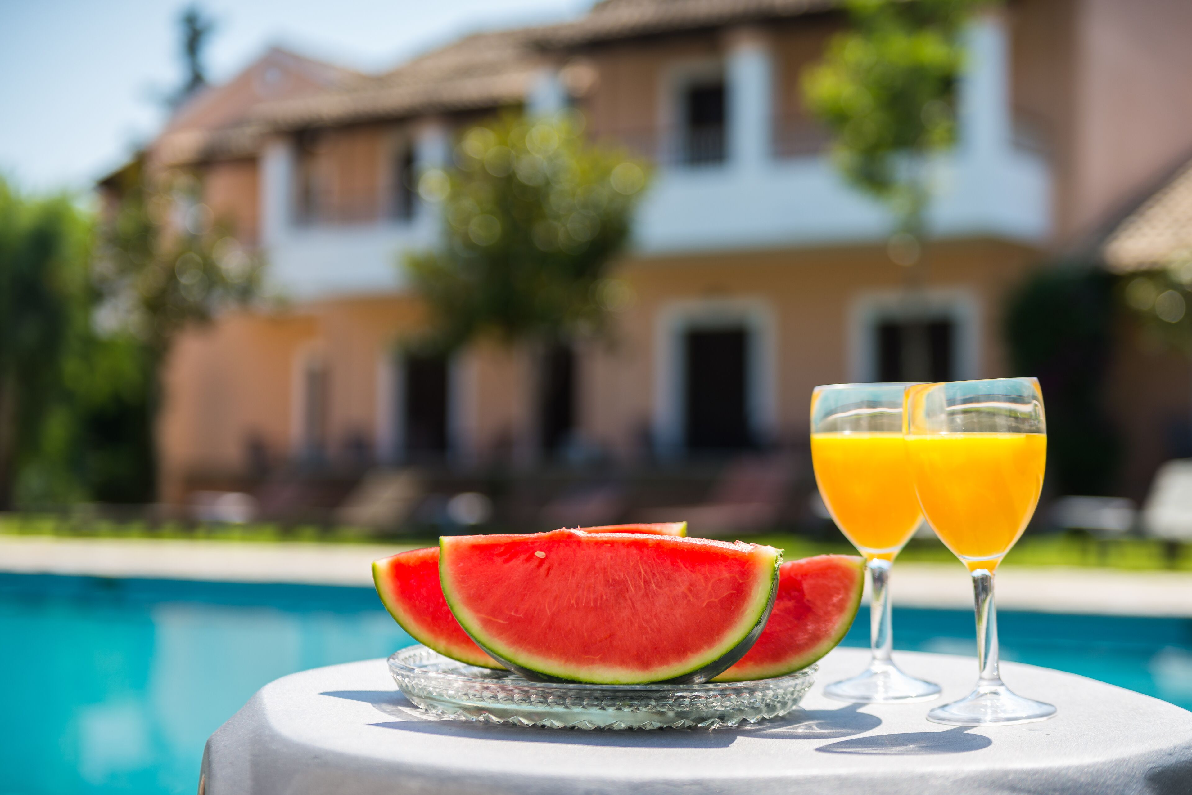A table with watermelon slices and two glasses of orange juice by a poolside, with a house in the background.