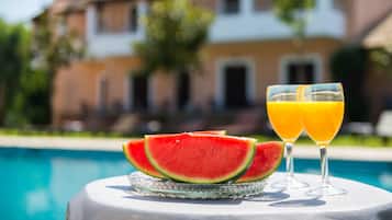A table with watermelon slices and two glasses of orange juice by a poolside, with a house in the background.