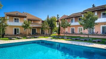 A swimming pool with lounge chairs, a clear blue sky, and a row of two-story buildings with balconies.