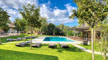 Una piscina rodeada de sillas de descanso y árboles, con un área de patio cubierta y un edificio al fondo.