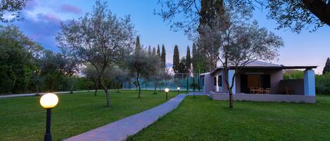 A pathway leading to a modern house with a glass facade, surrounded by trees and a well-maintained lawn.