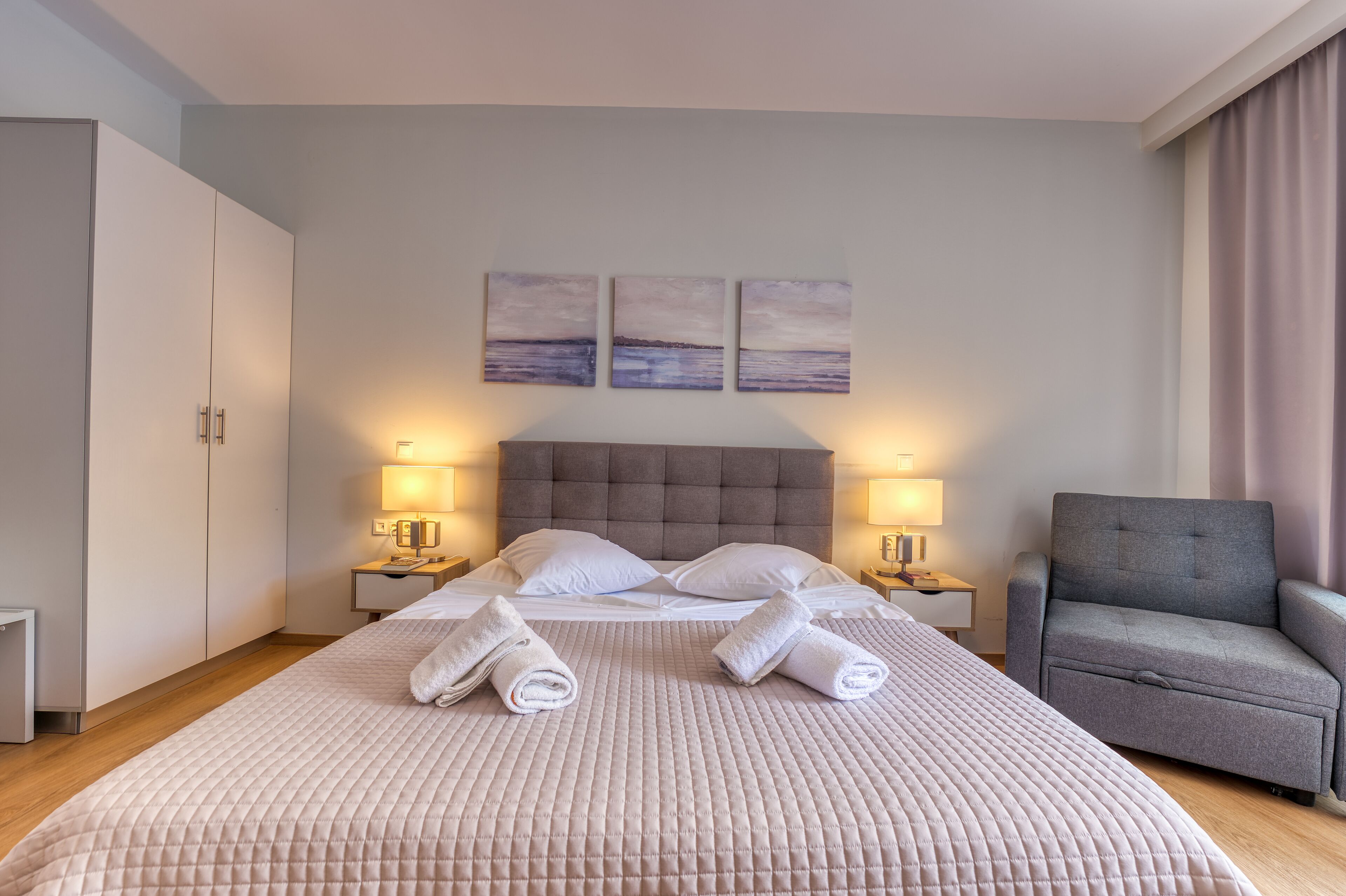 A neatly made bed with a beige quilt, two bedside lamps, and a grey armchair in a modern hotel room.