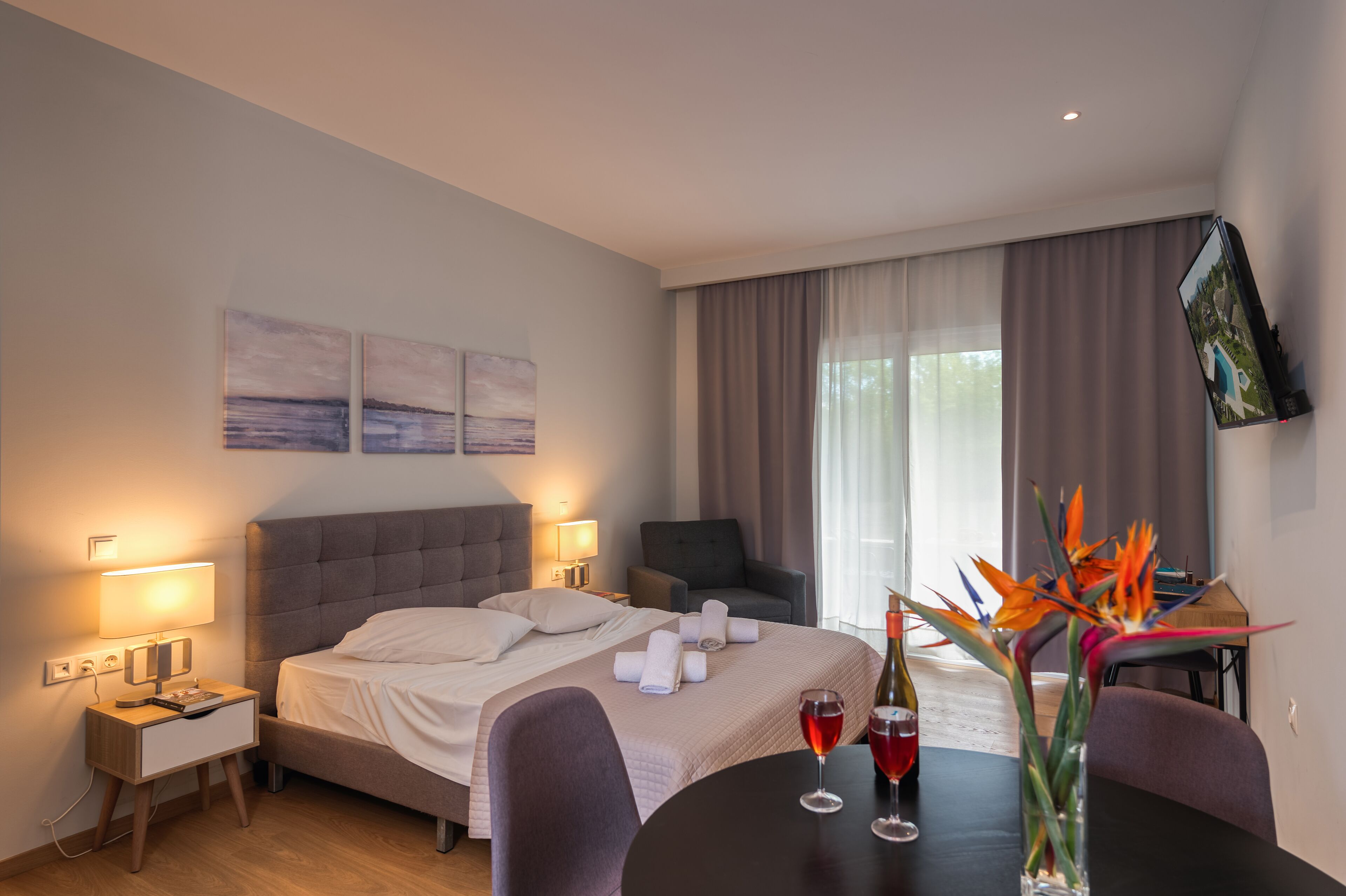 Une chambre d’hôtel comprenant un lit, des tables de chevet, un bureau, une chaise, une table à manger avec des verres à vin et un vase à fleurs.