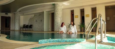 Una piscina interior con dos personas sentadas al borde, rodeada de paredes de azulejos y un diseño moderno.