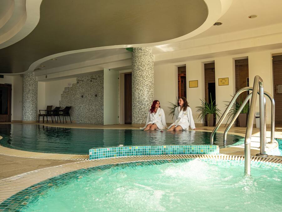 Una piscina interior con dos personas sentadas al borde, rodeada de paredes de azulejos y un diseño moderno.