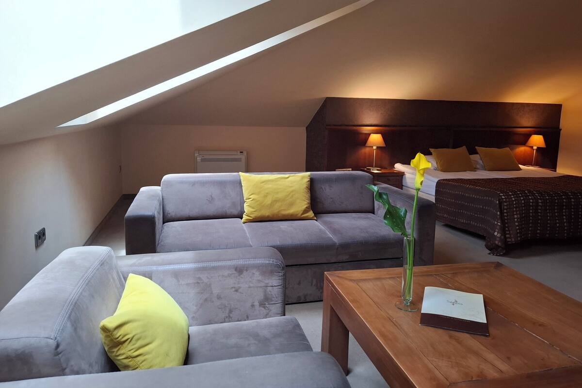 Ein modernes Schlafzimmer im Loft-Stil mit schrägem Dach, einer grauen Sofagarnitur, einem Holztisch und einem Bett mit dunkler Kopfteil.