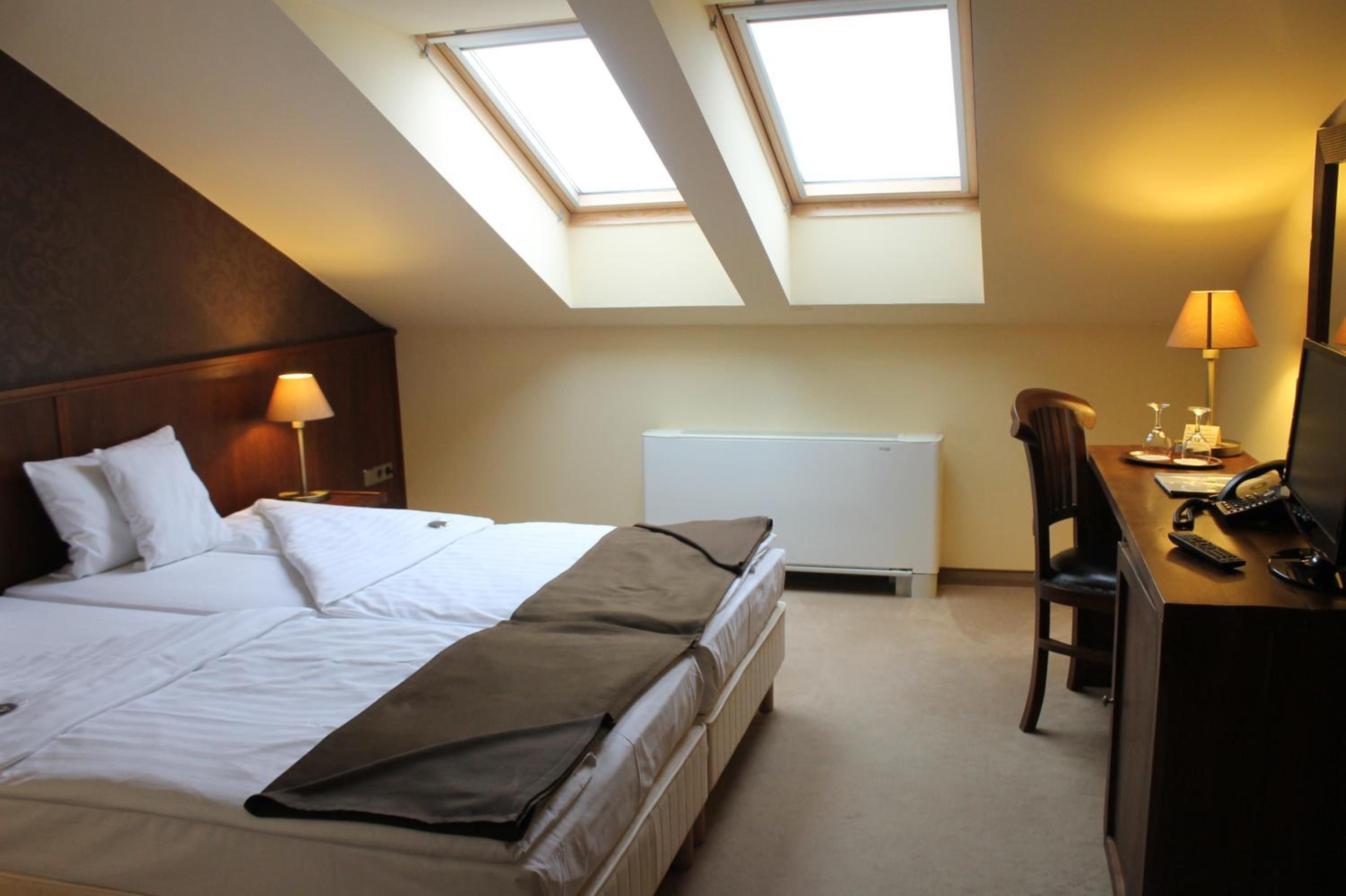 A hotel room with a bed, a desk, a chair, a radiator, and skylights.