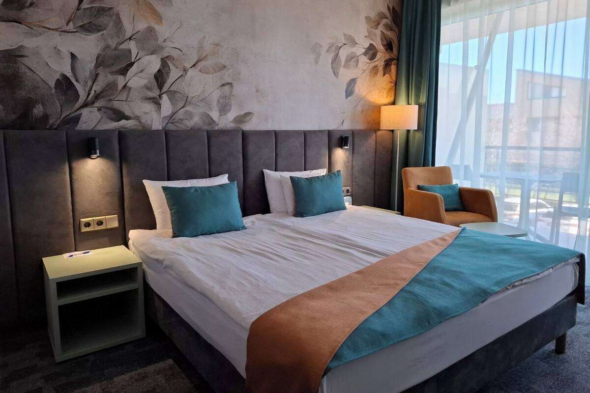 Ein Hotelzimmer mit einem großen Bett, zwei Nachttischen, einem Sessel und Blick ins Freie.