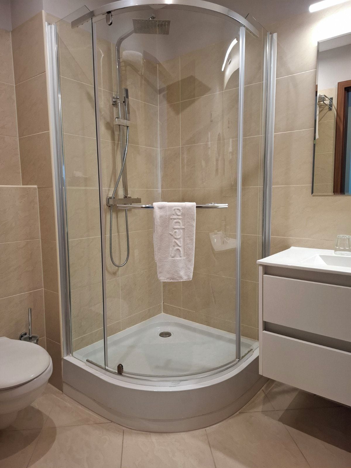 Eine begehbare Duschkabine mit Vorhang, ein Handtuch am Türgriff, ein weißes Waschbecken mit einem Glas und eine Toilette links.