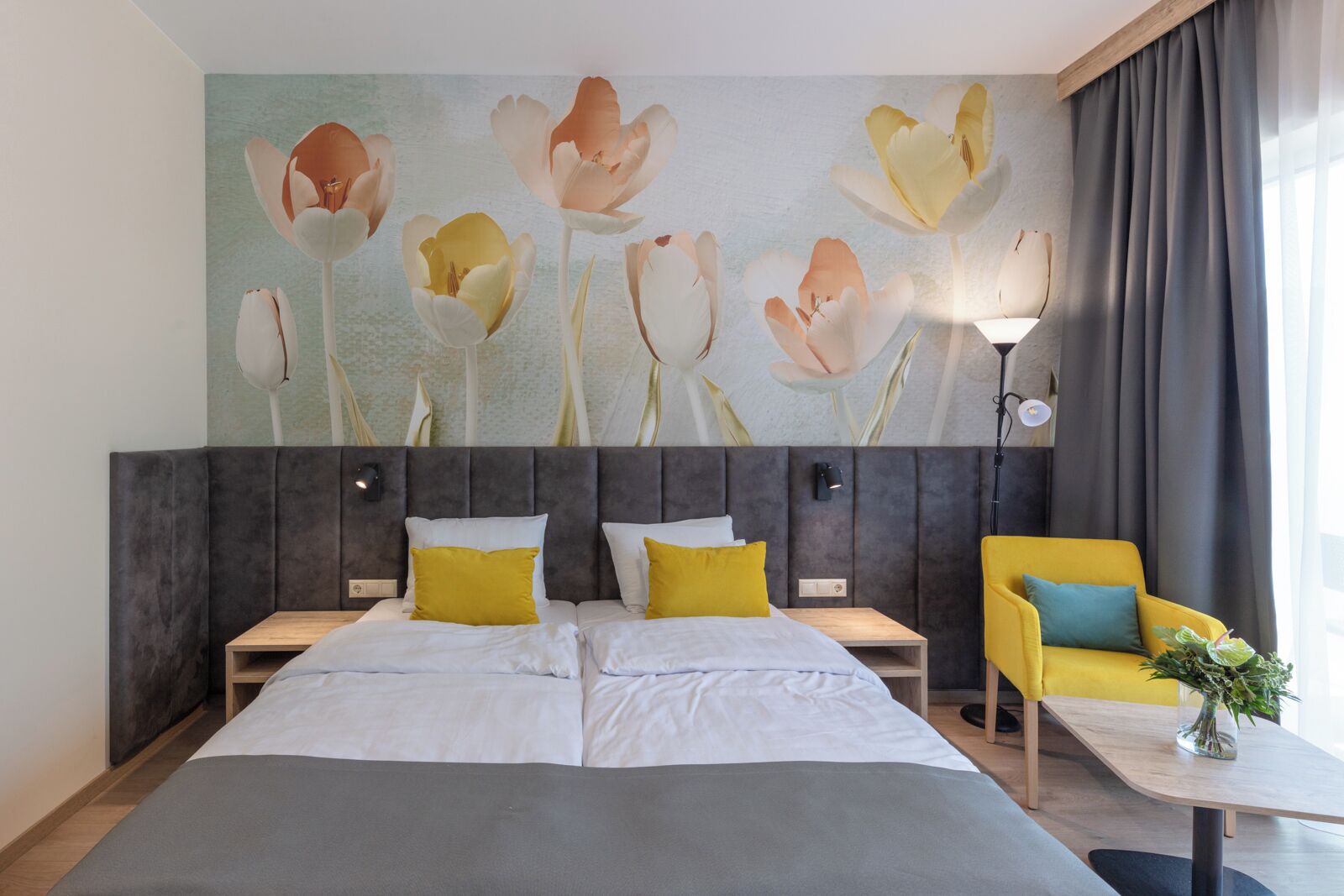 A hotel room with a large bed, two bedside tables, a nightstand, a yellow armchair, and a large floral wall mural.