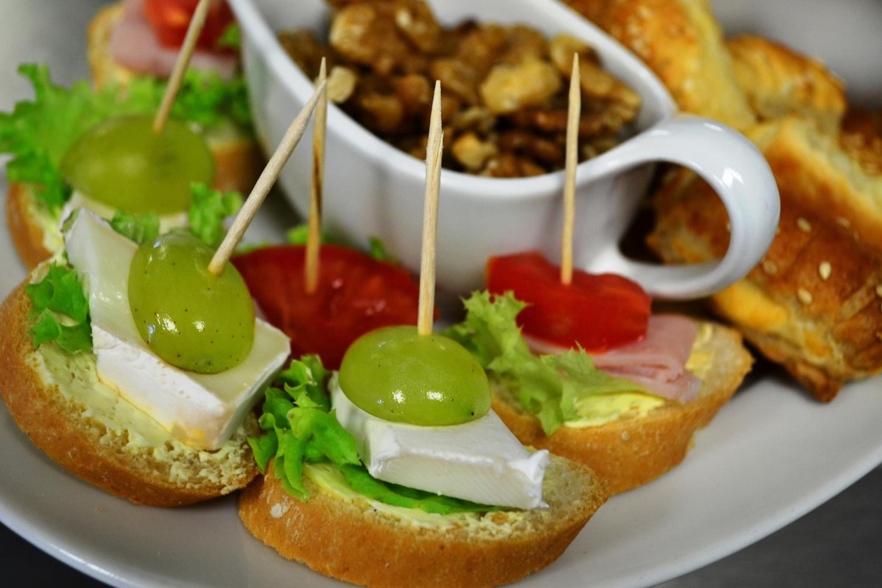 A plate with three open-faced sandwiches topped with cheese, lettuce, and a green grape, accompanied by a bowl of nuts and a slice of tomato.