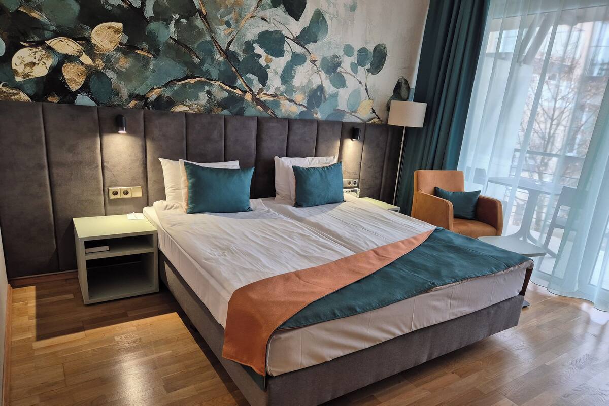 Ein modernes Hotelzimmer mit einem großen Bett, einem Nachttisch, einem Sessel und Blick ins Freie durch transparente Vorhänge.