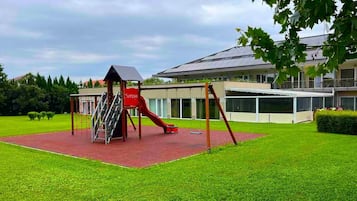 Un parque infantil con un tobogán y una estructura para escalar, rodeado de césped cuidado y un edificio con ventanas amplias al fondo.