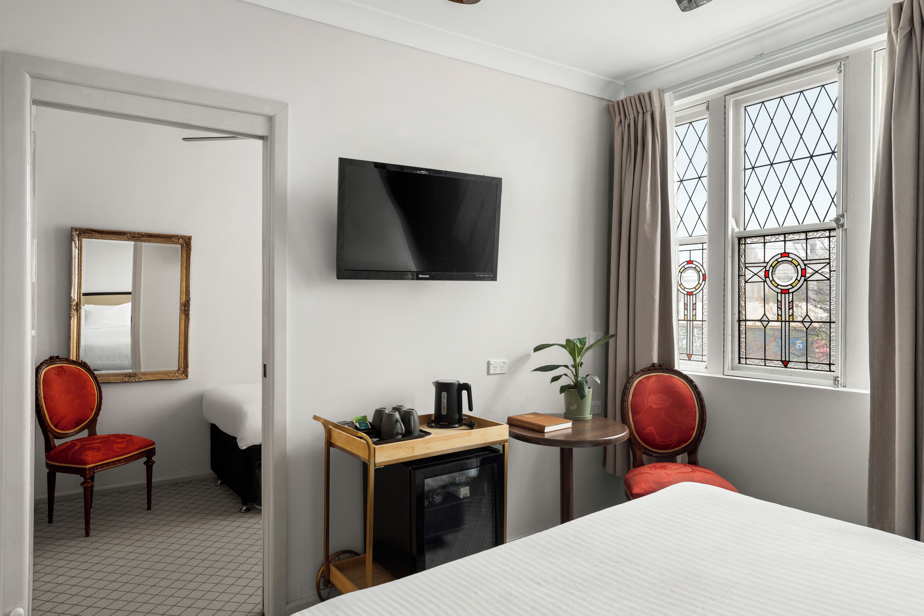 A hotel room with a bed, a TV, a small desk with a kettle, a chair, and a window with curtains.