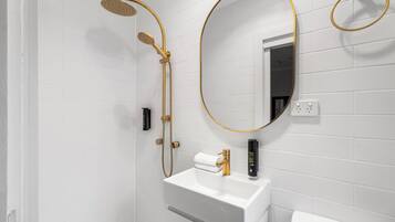A bathroom with a white tiled wall, a gold-framed mirror, a white sink, and a gold showerhead.