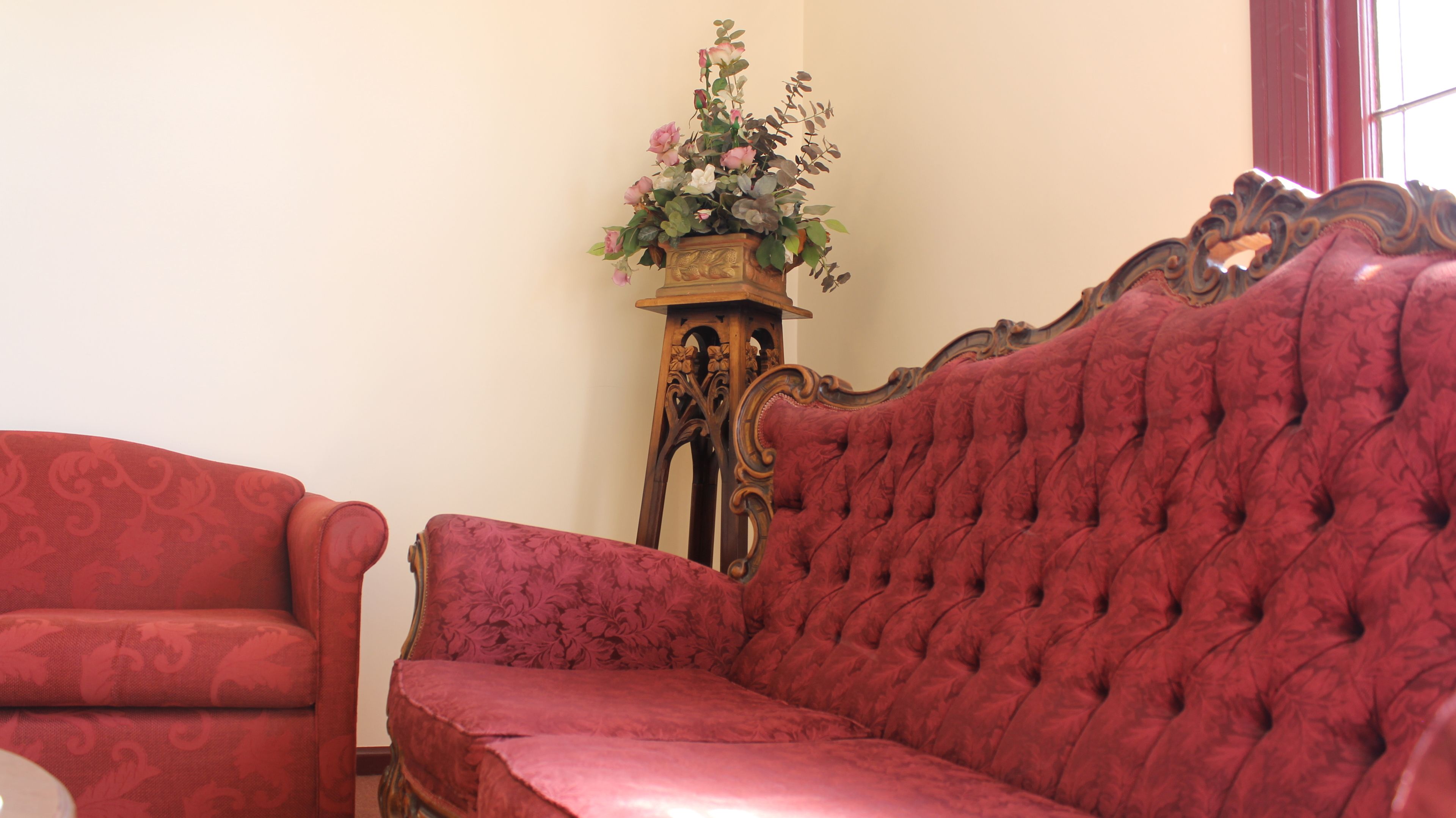 Ein Zimmer mit einer roten Stoffcouch und einem passenden Sessel, ein Holztisch mit einer Blumenkomposition und einem Fenster mit roten Vorhängen.