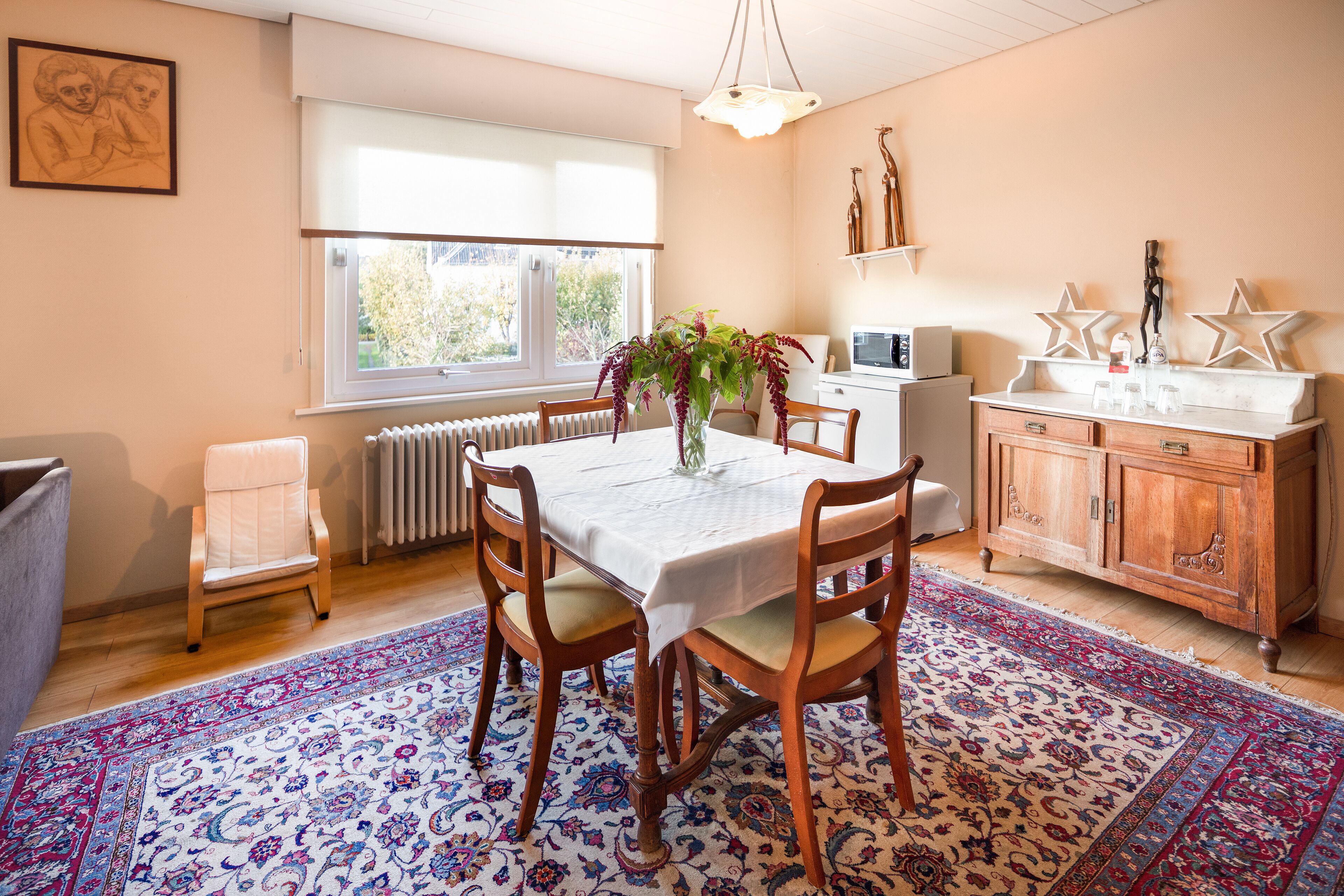 A dining area with a table set for four, surrounded by wooden chairs, a patterned rug, and a window with blinds.