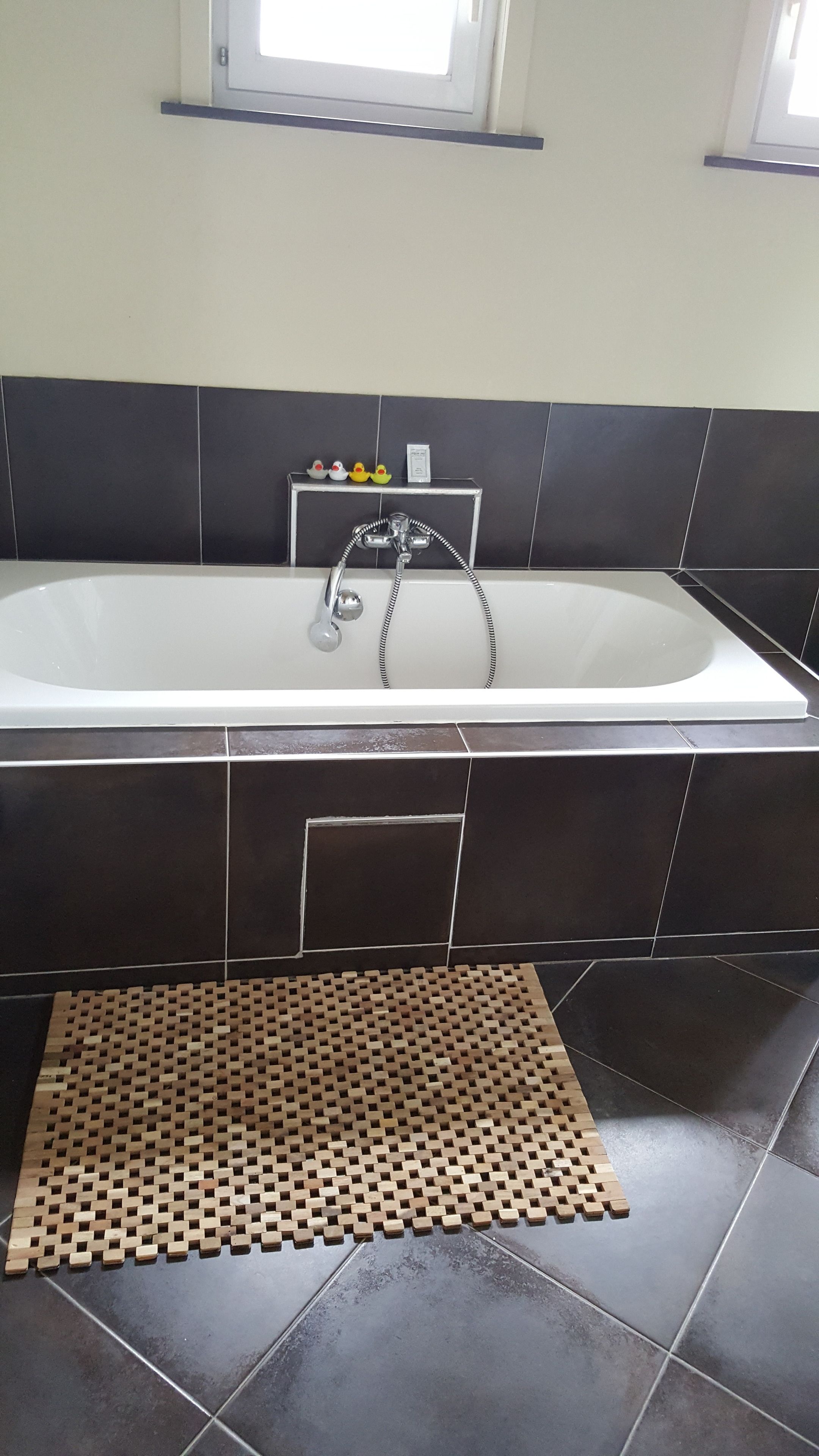 A bathroom with a bathtub, dark tiles, a wooden mat, and a window above the tub.