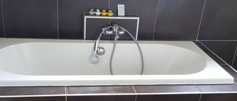 A bathroom with a bathtub, dark tiles, a wooden mat, and a window above the tub.