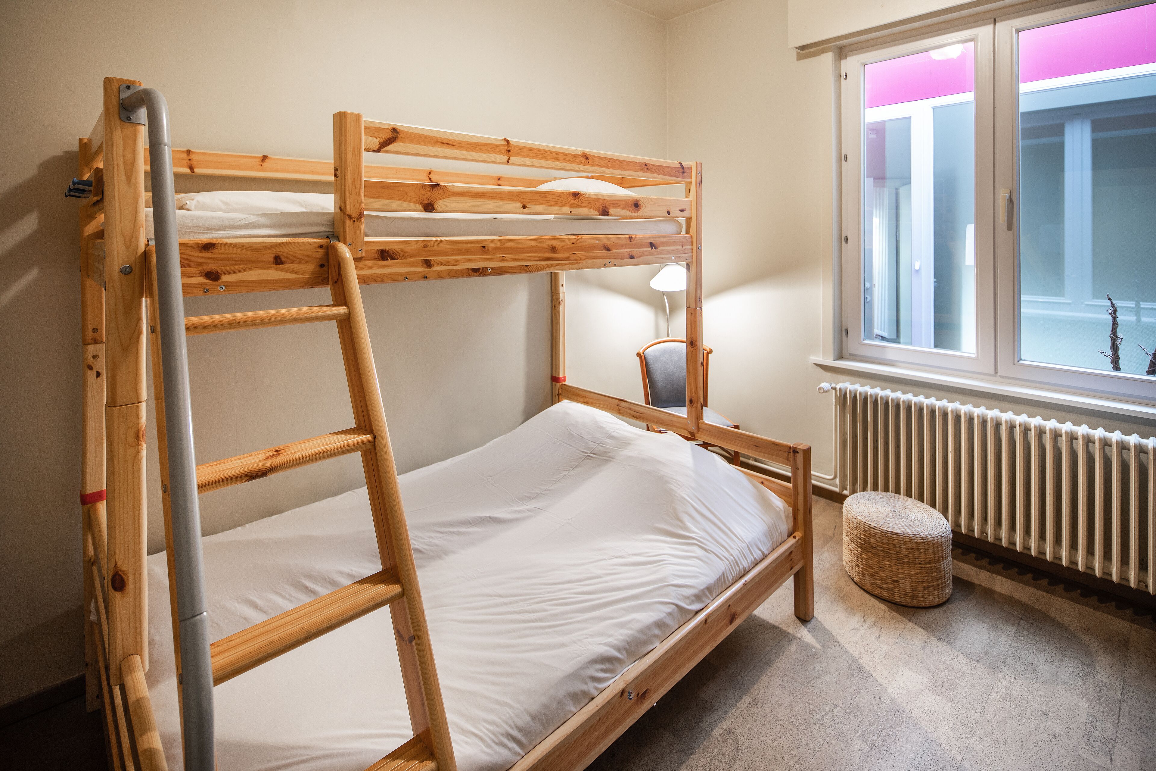 A bunk bed with a ladder, a chair, a small ottoman, and a window with a view of a building.