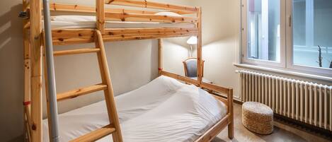 A bunk bed with a ladder, a chair, a small ottoman, and a window with a view of a building.